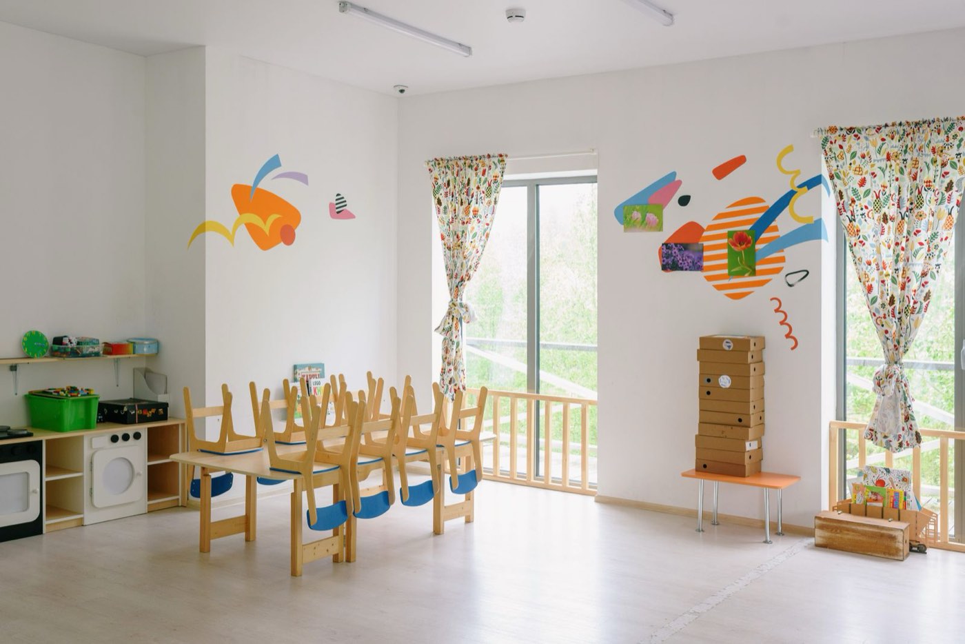 An empty nursery room at the end of the day: small wooden chairs turned up on a low table, a toy kitchen along the wall and bright light from a window.